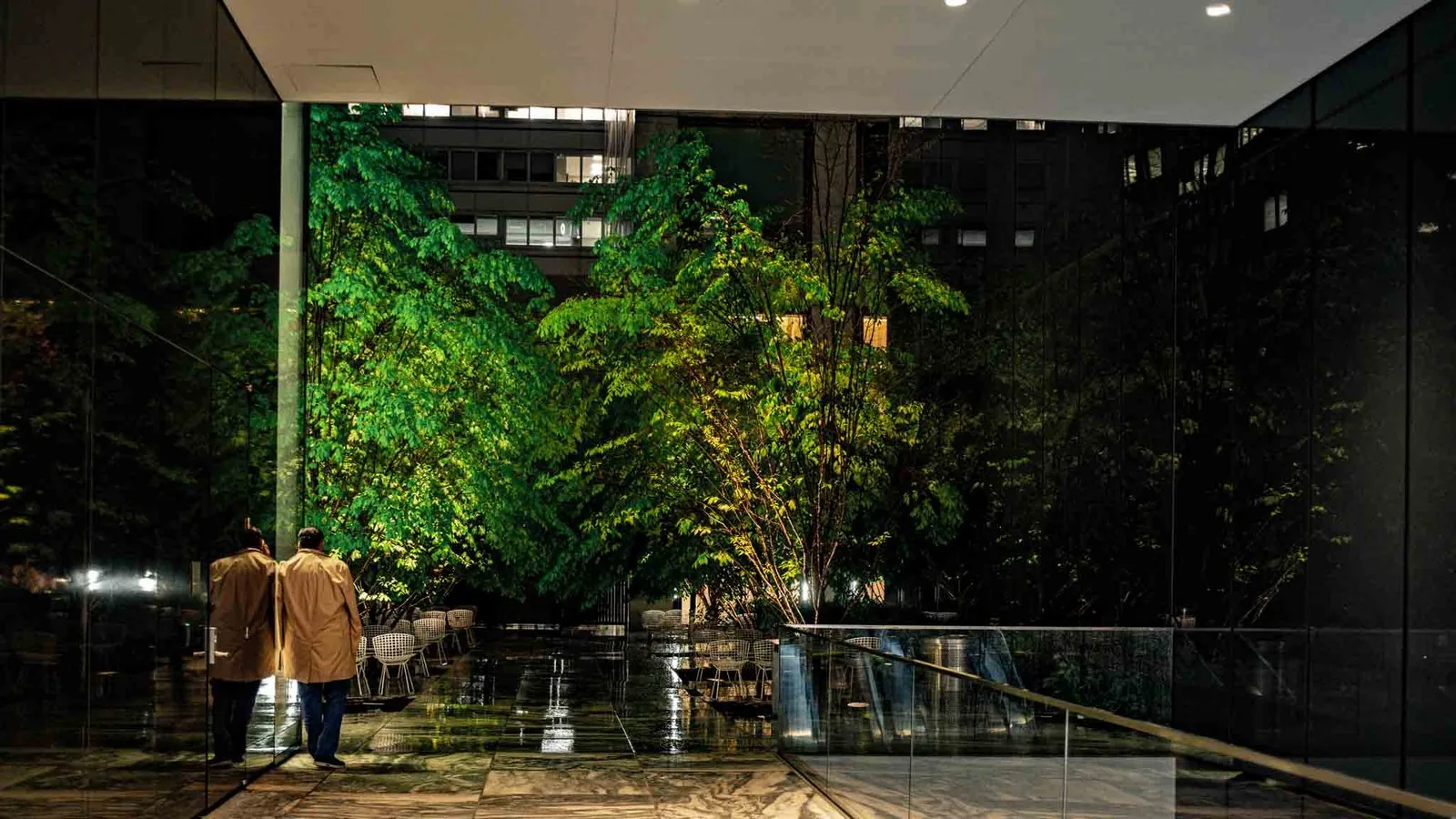 Two men in trench coats in the MoMA sculpture garden at night, lit trees and wet stone floor