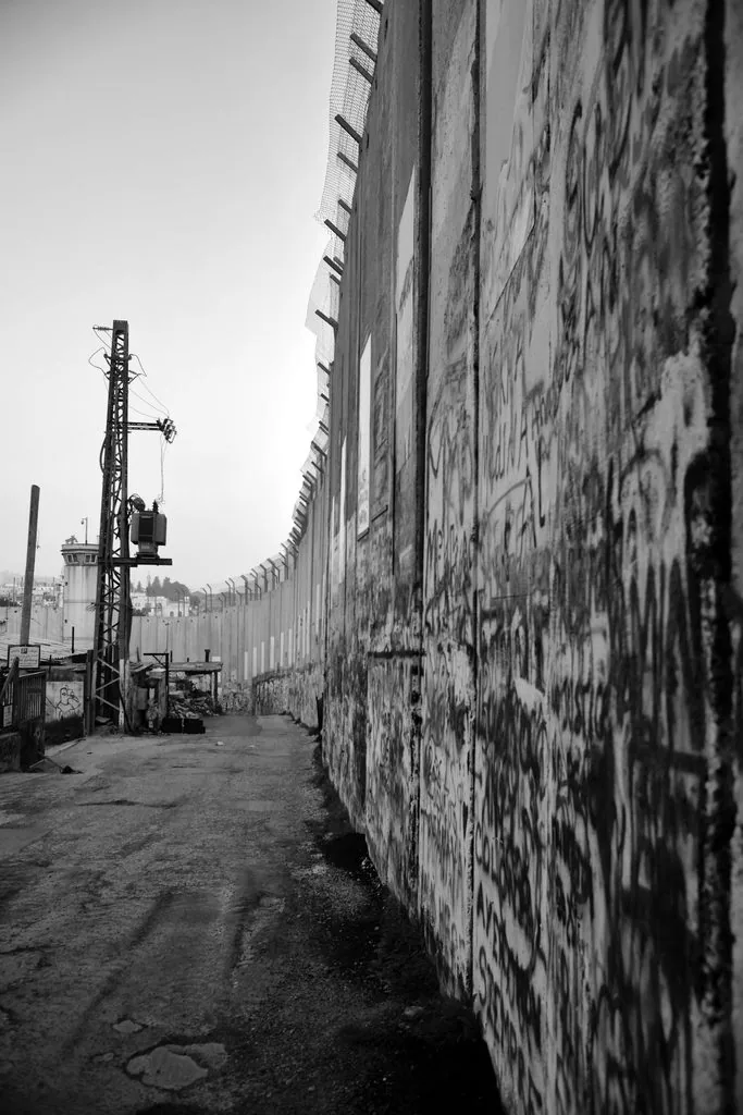 A long concrete wall covered in graffiti, a watchtower and power pylon at its far end, Holy Land