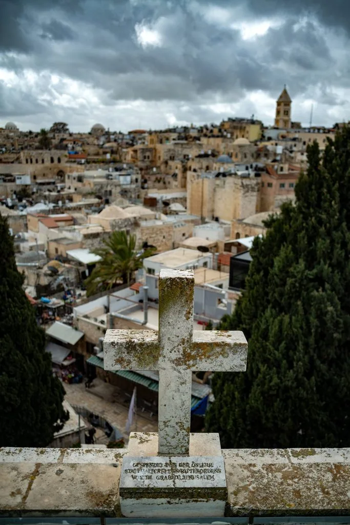 A weathered stone cross on a parapet above the rooftops of the Old City, a bell tower under heavy clouds, Old City, Jerusalem