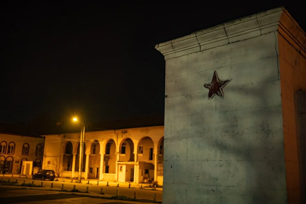 A whitewashed plinth with a red star at night, an arcaded building under a streetlamp behind it